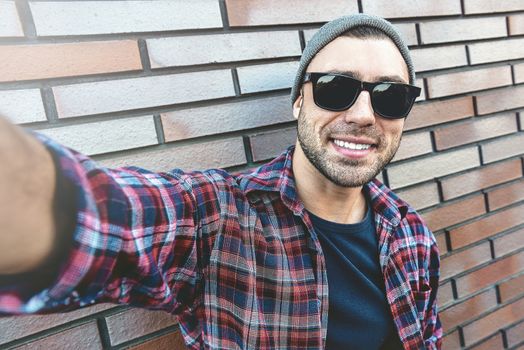 Portrait of stylish handsome young man with bristle standing outdoors and leaning on brick wall. Man wearing sunglasses and hat. Man making selfie.