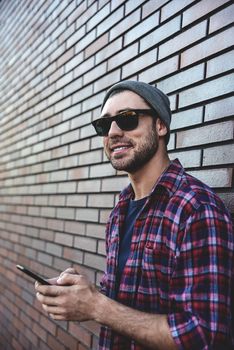 Hipster sms texting phone app in city street on brick wall background. Amazing man holding smartphone in smart casual wear standing. Urban young professional lifestyle.