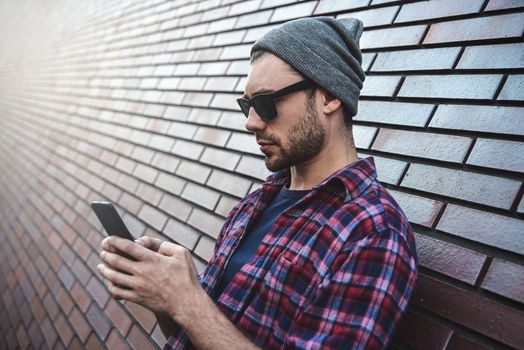 Hipster sms texting phone app in city street on brick wall background. Amazing man holding smartphone in smart casual wear standing. Urban young professional lifestyle.