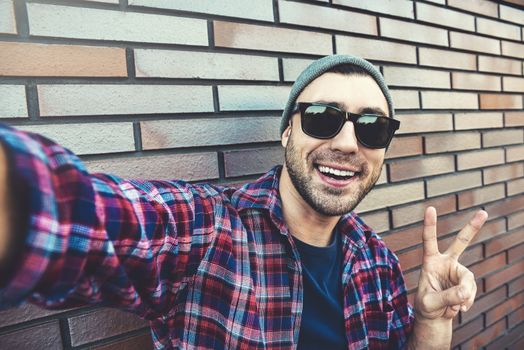 Portrait of stylish handsome young man with bristle standing outdoors and leaning on brick wall. Man wearing sunglasses and hat. Man making selfie.