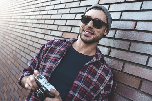 Smiling man with sunglasses hold retro photo camera Fashion Travel Lifestyle outdoor while standing against brick wall background.