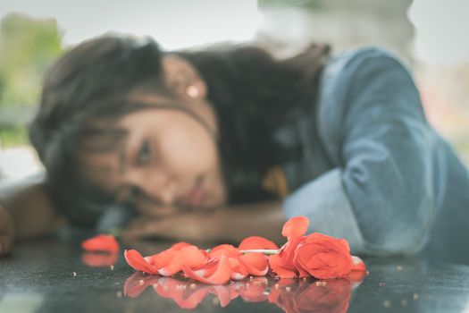 Selective focus on red rose petals, lonely young teenager sitting sadly on table by laying her head down - concept of love breakup or broken heart