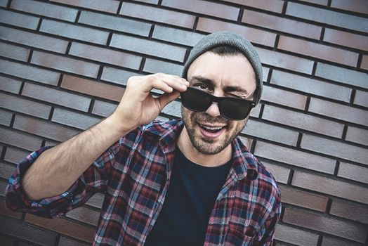 Young adult man wearing sunglasses standing over brown brick wall smiling with happy face winking at the camera.