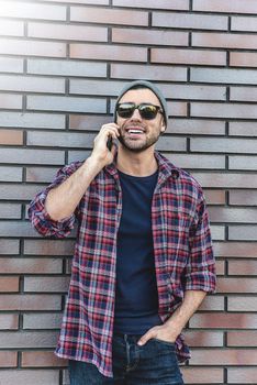 Hipster calling in city street on brick wall background. Amazing man holding smartphone in smart casual wear standing. Urban young professional lifestyle.