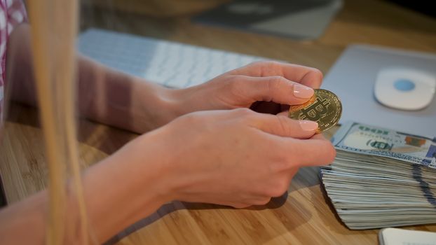 Crop view of hands of woman holding shiny metal bitcoin sitting at wooden desk with wad of cash nearby at night