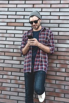 Hipster sms texting phone app in city street on brick wall background. Amazing man holding smartphone in smart casual wear standing. Urban young professional lifestyle.