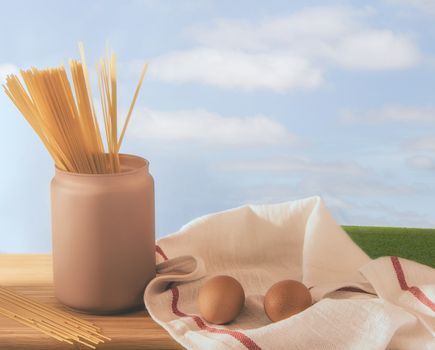 Gourmet still life with a pink spaghetti jar and two eggs in kitchen towels on a wooden board and a meadow at the end with a cloudy sky. Light vintage effect. Cooking pasta on a farm. Copy space.