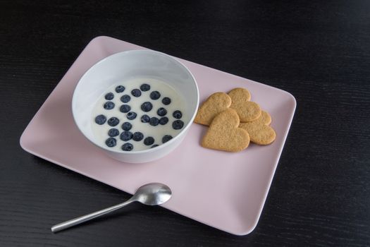 Breakfast. Yogurt with blueberries heart shaped cookies. Ingredients in a pink tray on a black wood table. Copy space.