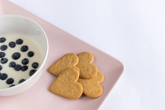 Breakfast. Yogurt with blueberries heart shaped cookies. Ingredients in a pink tray on a white background. Detail from above with copy space.