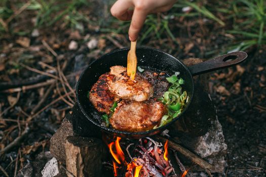 Frying meat in a pan over an open fire with leek. Steak in a pan on a fire. Cooking in nature. Picnic. Grill on fire. Hand with a wooden spatula.