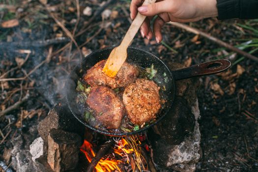 Frying meat in a pan over an open fire with leek. Steak in a pan on a fire. Cooking in nature. Picnic. Grill on fire. Hand with a wooden spatula.