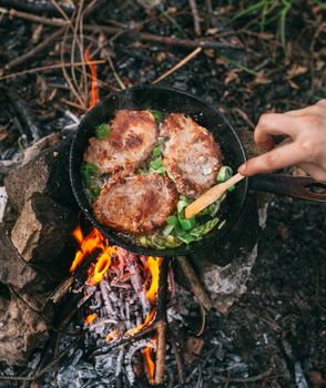 Frying meat in a pan over an open fire with leek. Steak in a pan on a fire. Cooking in nature. Picnic. Grill on fire. Hand with a wooden spatula.