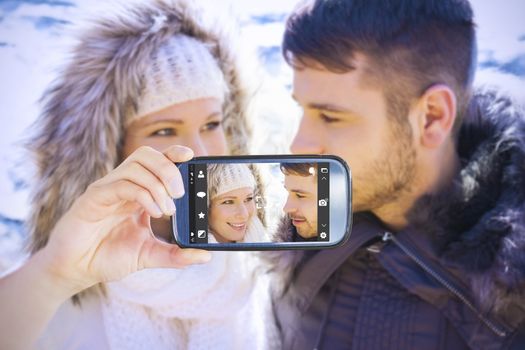Hand holding smartphone showing against couple in fur hood jackets against snowed mountain