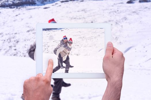 Hand holding tablet pc against full length of a man piggybacking cheerful woman on snow