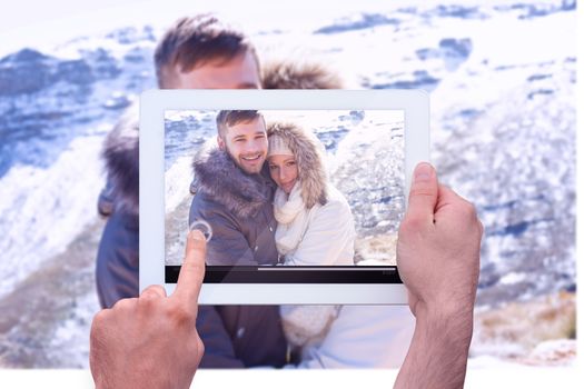 Hand holding tablet pc against couple in jackets embracing against snowed mountain