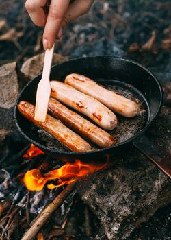 Roasting sausages in a frying pan over an open fire. Preparing food in nature. Lunch in the open air. Hand with a wooden shovel. Picnic in the forest. Food on fire