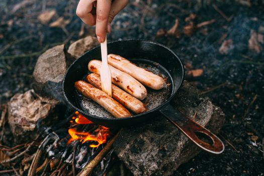 Roasting sausages in a frying pan over an open fire. Preparing food in nature. Lunch in the open air. Hand with a wooden shovel. Picnic in the forest. Food on fire