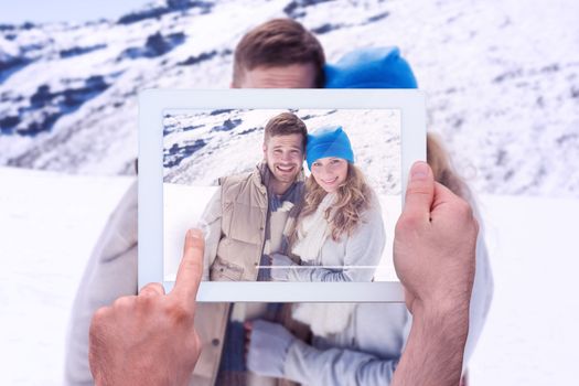 Hand holding tablet pc against loving couple in warm clothing on snow covered landscape
