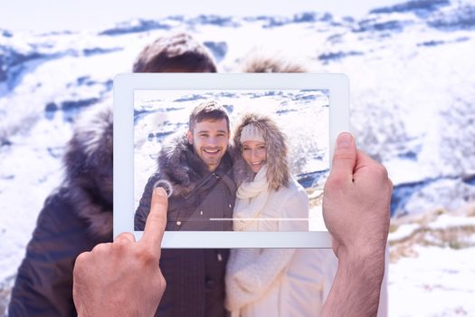 Hand holding tablet pc against couple in fur hood jackets against snowed mountain range