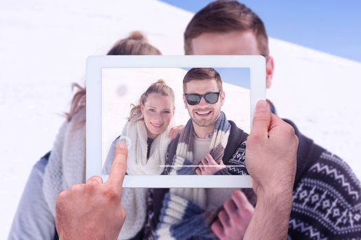 Hand holding tablet pc against smiling couple in warm clothing on snow covered landscape