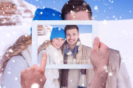 Hand holding tablet pc against couple in warm clothing on snow covered landscape