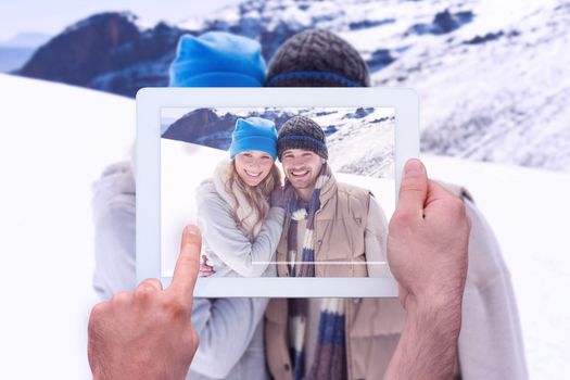 Hand holding tablet pc against couple in warm clothing on snow covered landscape