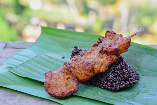Thai street food. Delicious Chicken mixed with Thai herb grilled with wooden stick served with black sticky rice on green banana leave. Reduce plastic for save earth.
