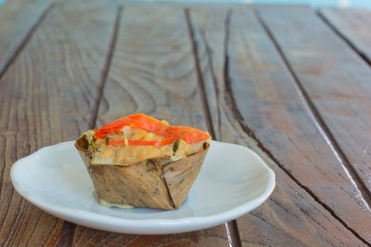 Thai steamed seafood curry in cup made from dry banana leave on wooden table and red chilli topping. Delicious Thai food.