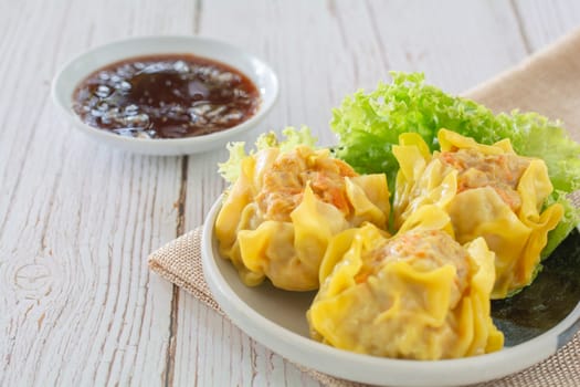 Chinese Steamed Dumpling, Shumai on white dish served with soy sauce and 
lettuce leaves on brown cloth and wooden table. Delicious Dimsum pork.