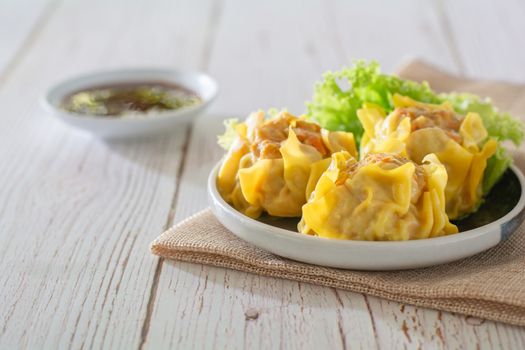 Chinese Steamed Dumpling, Shumai on white dish served with soy sauce and 
lettuce leaves on brown cloth and wooden table. Delicious Dimsum pork.