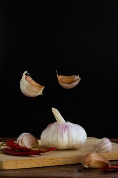 Group of garlic on chopping board and some garlic cloves floating in the air and red dried chilli on wooden table with black background. Copy space for your text.