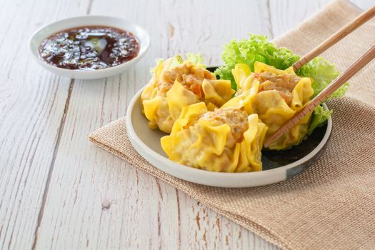 Chinese Steamed Dumpling, Shumai on white dish served with soy sauce and 
lettuce leaves on brown cloth and wooden table. Delicious Dimsum pork.