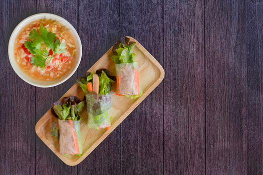 Top view fresh organic green vegetable (lettuce and carrot slice) rice wraps with tuna, sausage on wooden dish and Thai seafood dipping sauce in white bowl on dark wooden background. Healthy Thai food for loose weight. 