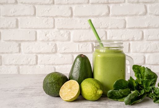 Avocado, lime and spinach smoothie in mason jar decorated with ingredients front view on white brick wall background
