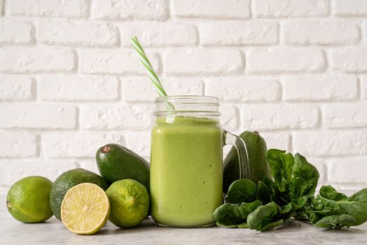Avocado, lime and spinach smoothie in mason jar decorated with ingredients front view on white brick wall background