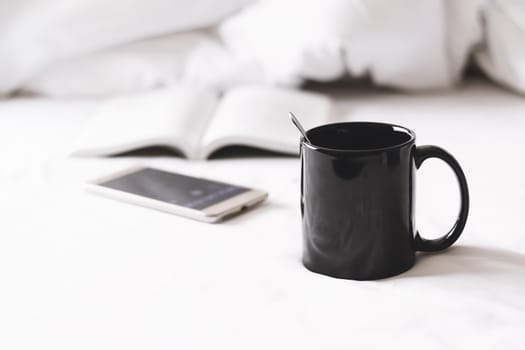 a coffee cup with smartphone and a book on white bed