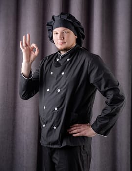 Young smiling male chef showing ok sign isolated on dark curtain background