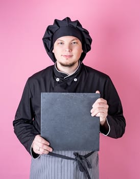 Young smiling male chef holding a square board isolated on pink background with copy space