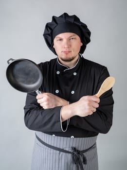 frowning male chef in black uniform holding a saucepan and a spoon isolated on gray background