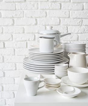 piles of white ceramic dishes and tableware on the table on white brick wall background with copy space