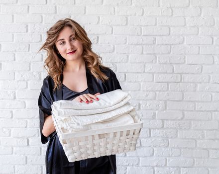 Laundry concept. Young woman holding a white basket with towels on white bricks background with copy space