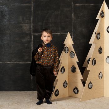 on a dark background little boy posing in a stylish costume near a Christmas tree made of wood