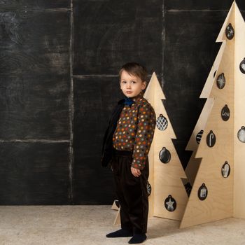 on a dark background little boy posing in a stylish costume near a Christmas tree made of wood