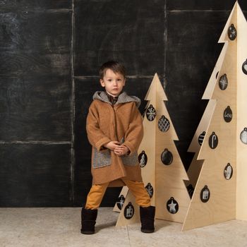 on a dark background little boy posing in a stylish coat near a Christmas tree made of wood