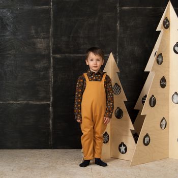 on a dark background little boy posing in a fashionable costume near a Christmas tree made of wood