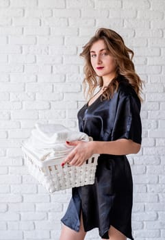 Laundry concept. Young attractive woman holding a white basket with towels on white bricks background