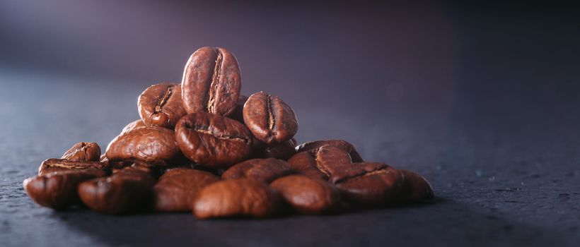 Grains of black coffee on a black granite countertop with yellow light from behind. Advertising photo of coffee. Photo with a glare from the light. Back light on a pyramid with grains.