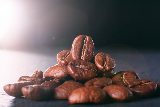 Grains of black coffee on a black granite countertop with yellow light from behind. Advertising photo of coffee. Photo with a glare from the light. Back light on a pyramid with grains.