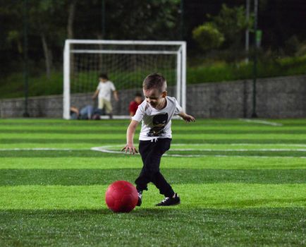 cheerful boy playing with red ball on the background of a football goal, in the twilight time