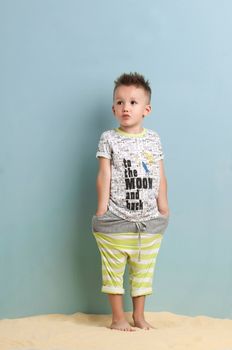 little boy in a shirt and shorts standing on the sand on a light blue background